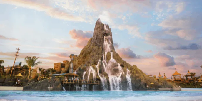 A wide shot of a massive artificial volcano with cascading waterfalls flowing into a large, wavy blue pool, under a pastel pink and blue sunset sky with scattered clouds